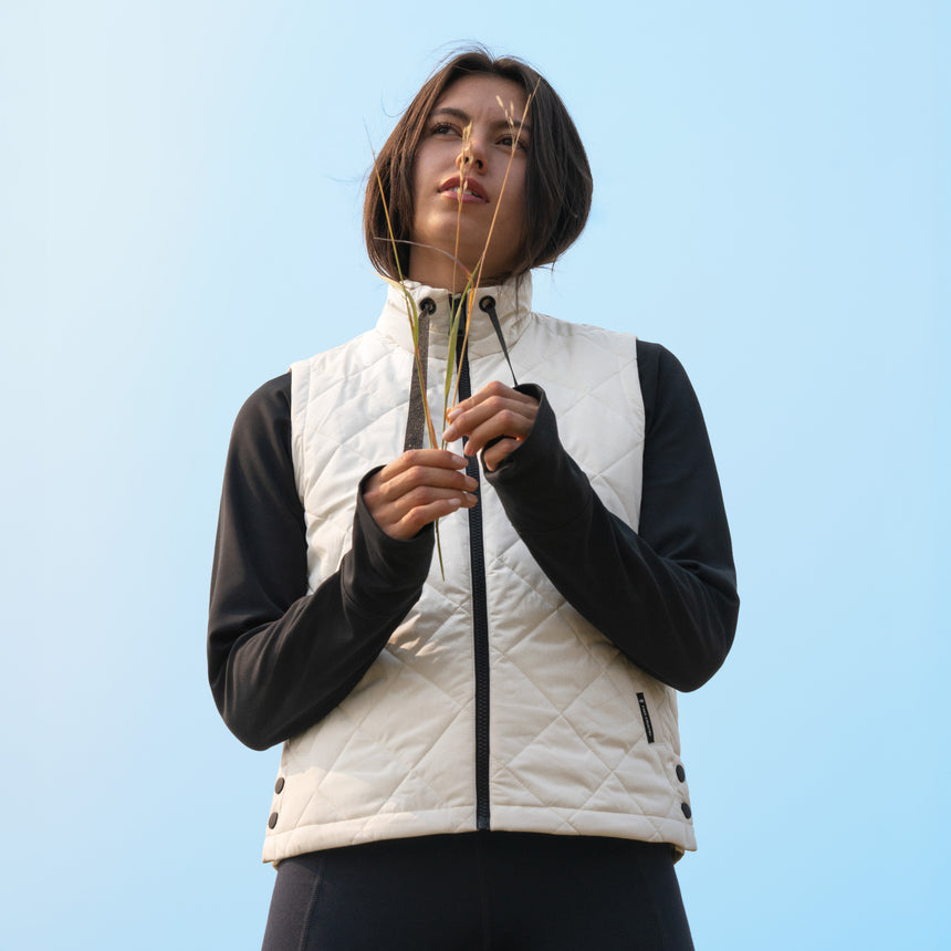 A woman wearing a white Free Country Women’s Cirrus Diamond Quilted Vest over a black long-sleeved shirt, holding a sprig of grass against a light blue sky.