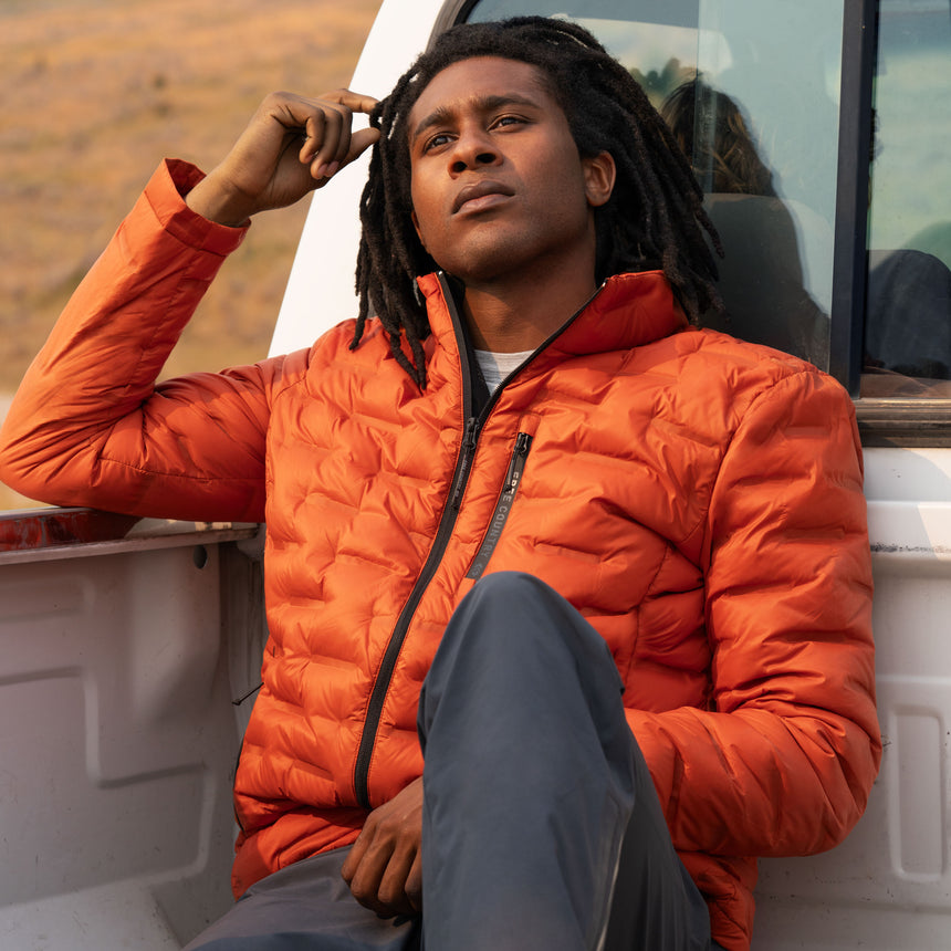 A man wearing a lightweight, packable orange Men’s Paramount Down Packable Puffer Jacket by Free Country sits in the bed of a pickup truck.
