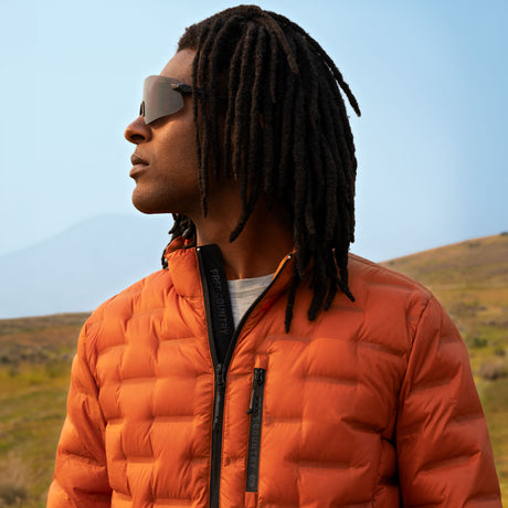 A man wearing a rust-colored Men’s Paramount Down Packable Puffer Jacket with a zippered front, viewed from the side outdoors.