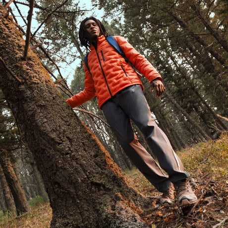 A person wearing an orange Men’s Paramount Down Packable Puffer Jacket by Free Country and dark trousers leans against a tree in a forest.