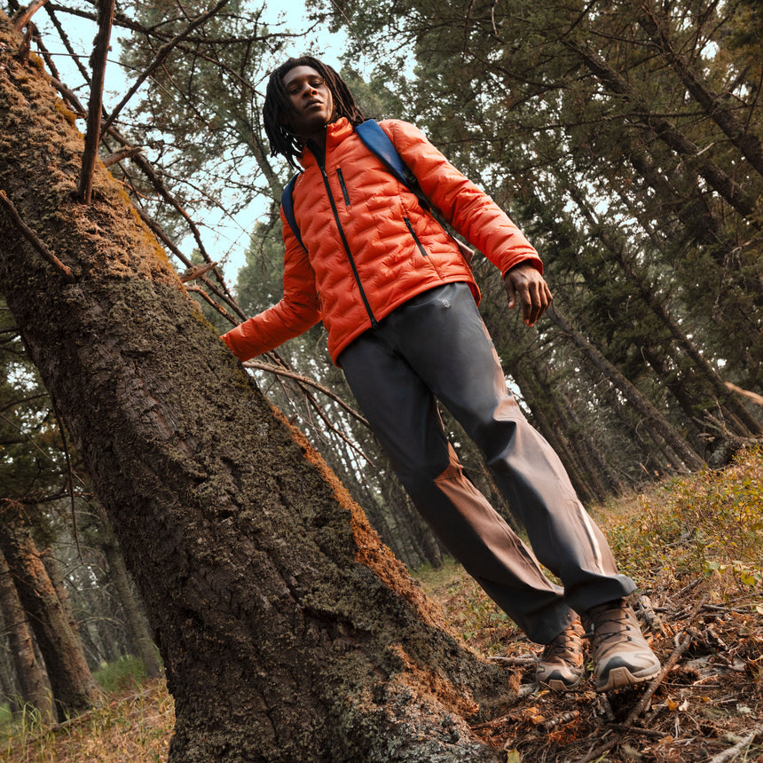 A person wearing an orange Men’s Paramount Down Packable Puffer Jacket by Free Country and dark trousers leans against a tree in a forest.