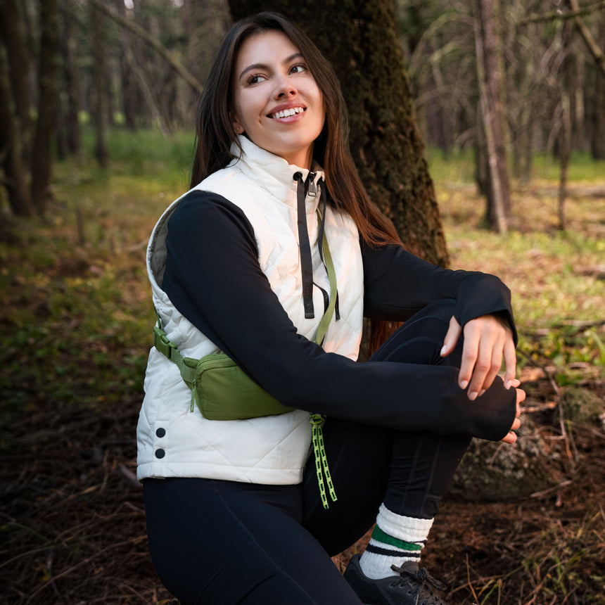A woman wearing a white Free Country Women’s Cirrus Diamond Quilted Vest, black long-sleeved shirt, and a green crossbody bag crouches in a wooded area.
