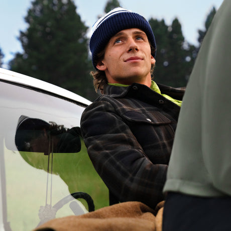 A man wearing a plaid Free Country Men’s Chill Out Fleece Shirt Jacket and a striped knit beanie looks upward while leaning against a vehicle.