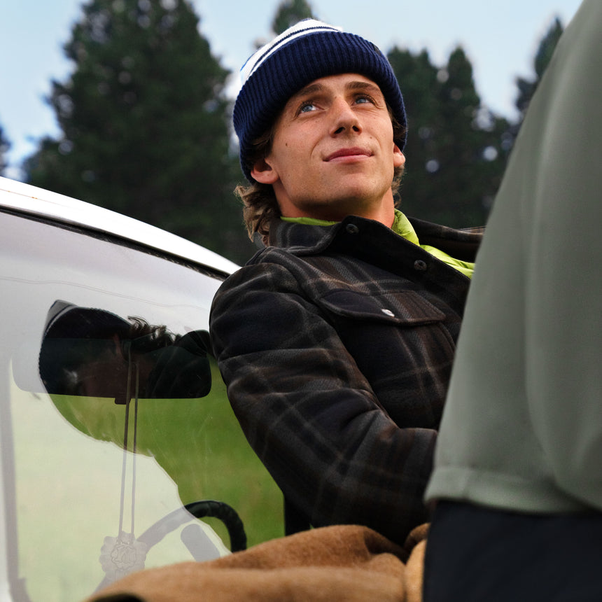 A man wearing a plaid Free Country Men’s Chill Out Fleece Shirt Jacket and a striped knit beanie looks upward while leaning against a vehicle.