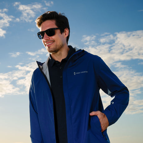A man smiles outdoors with one hand in his pocket, wearing sunglasses and a Free Country Men's Hydro Lite X2O Spectator Waterproof Jacket against a blue sky with scattered clouds.