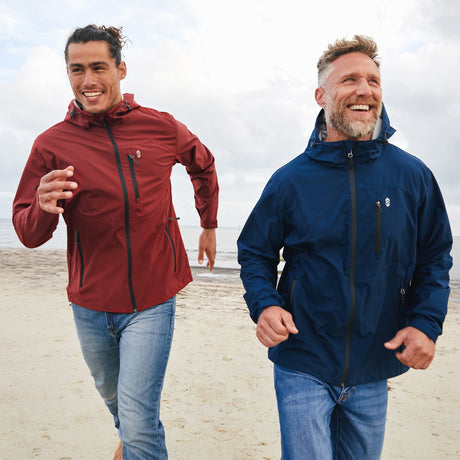 Two men wearing Clearance Men's Hydro Lite X2O Highline Waterproof Jackets—one in red, one in navy—run and smile on a sandy beach under cloudy skies. Dressed in blue jeans, they appear happy and active.