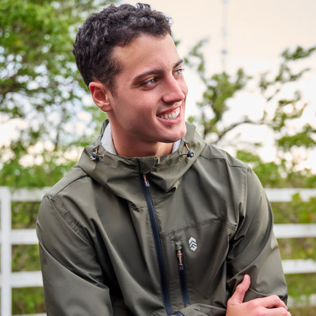A young man smiles outdoors, wearing the Clearance Men's Hydro Lite X2O Highline Waterproof Jacket. Green trees and a white fence are behind him under a slightly cloudy sky.