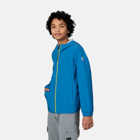 A smiling child with curly hair wears the Free Country Boys' Hydro Lite Recess X2O Rain Jacket and light gray pants, standing with one hand in their pocket against a plain white background.