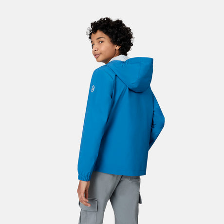 A boy with short curly hair wearing the Free Country Boys' Hydro Lite Recess X2O Rain Jacket stands facing away, turning slightly to look over his shoulder against a plain light background.