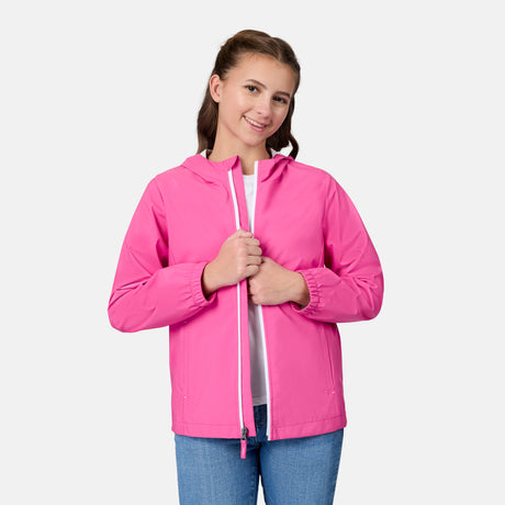 A young person with long brown hair smiles at the camera, wearing a bright pink Free Country Girls' Recess X2O Rain Jacket over a white shirt and blue jeans, standing against a plain light background.