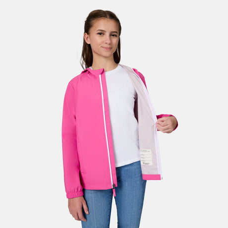 A girl smiles, wearing the Free Country Girls' Recess X2O Rain Jacket in bright pink over a white t-shirt and blue jeans. She holds the lightweight coat open to show its light lining, standing against a plain white background.