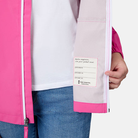 A girl in blue jeans opens her Free Country Girls' Recess X2O Rain Jacket, showing a label inside that reads, "Hello explorer, I’m your jacket now!" with spots for name and contact info.