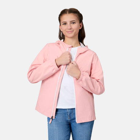 A young woman with braided brown hair smiles as she zips up a light pink Free Country Girls' Recess X2O Rain Jacket over a white shirt and blue jeans, standing against a plain white background.