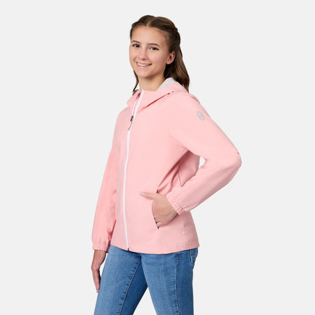 A young woman with long brown hair, wearing a light pink Free Country Girls' Recess X2O Rain Jacket and blue jeans, stands smiling with one hand in her pocket against a plain white background.