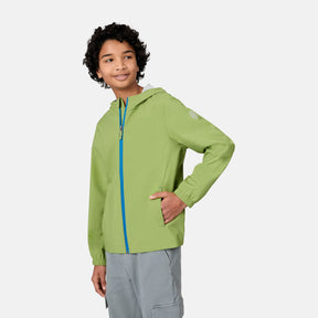 A young person with curly hair wears the Free Country Boys' Hydro Lite Recess X2O Rain Jacket in light green with a blue zipper and gray pants, standing against a plain light background, one hand in their pocket, looking to the side and smiling.