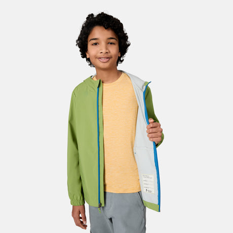 A smiling child with curly hair models the Free Country Boys' Hydro Lite Recess X2O Rain Jacket, worn over a yellow shirt and gray pants, standing against a plain light background and holding the jacket open to reveal its interior.