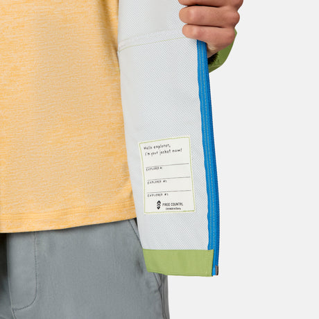A person holds open the light gray sleeve of the Free Country Boys' Hydro Lite Recess X2O Rain Jacket, which has a blue zipper, green cuff, and an inside label that reads "Hello explorer, I'm your jacket now!" with Explorer # and a recycling symbol.