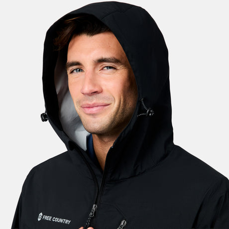 A man with short brown hair smiles slightly while wearing the Free Country Men's Hydro Lite X2O Status Jacket in black with the hood up. The background is plain white.