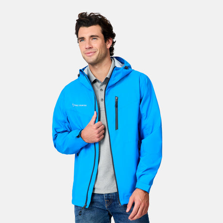 A man smiles as he stands against a plain light background, wearing the Free Country Men's Hydro Lite X2O Status Jacket over a gray polo and blue jeans, holding the front of his lightweight rain jacket with one hand.