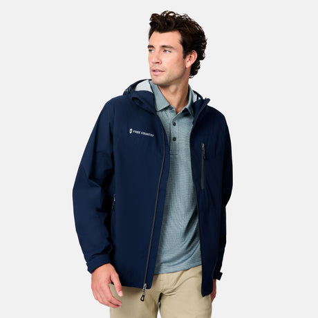 A man models the Free Country Men's Hydro Lite X2O Status Jacket in navy blue over a light blue patterned polo and beige pants, standing against a plain light gray background and looking to his left.