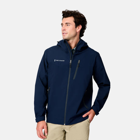 A man stands against a light gray background, looking slightly left, wearing khaki pants and the Free Country Men's Hydro Lite X2O Status Jacket—a navy blue, lightweight, hooded rain jacket with a chest pocket and brand logo.