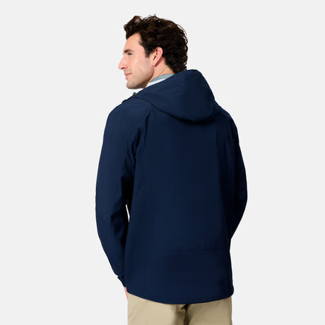 A man faces away from the camera wearing a Free Country Men's Hydro Lite X2O Status Jacket in navy blue and beige pants, standing against a plain light background.