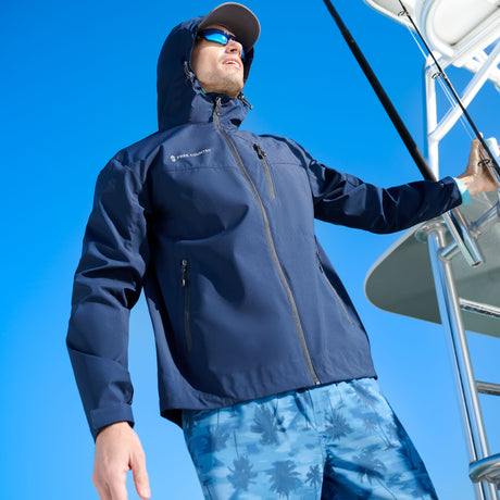 A man in the Free Country Men's Hydro Lite X2O Status Jacket, blue patterned shorts, sunglasses, and a cap stands on a boat holding the railing with a clear blue sky behind him.