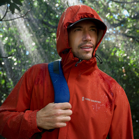 A person wearing the Free Country Men's Hydro Lite X2O Spectator Waterproof Jacket and a black cap stands in a green forest, holding a blue-strap backpack as sunlight filters through the trees and raindrops shimmer on their jacket.
