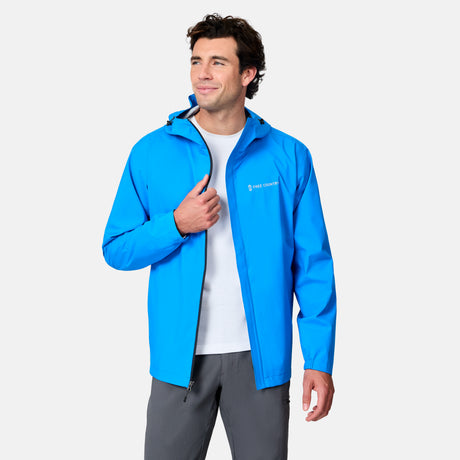 A man wearing the Free Country Men's Hydro Lite X2O Spectator Waterproof Jacket in bright blue over a white shirt and gray pants stands against a light background, holding the jacket’s edge and smiling slightly while looking to the side.