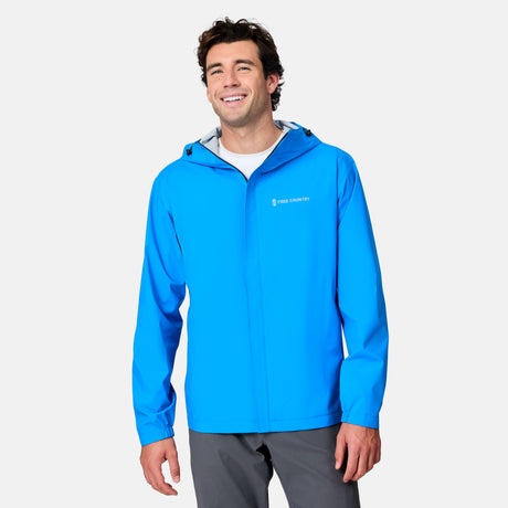A smiling man in a bright blue Free Country Men's Hydro Lite X2O Spectator Waterproof Jacket and gray pants stands against a white background. The lightweight jacket features a zipper and waterproof protection.