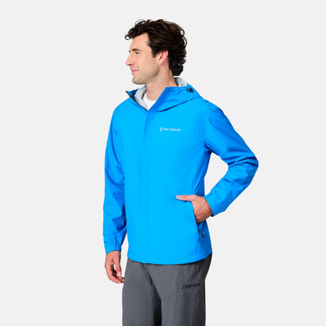 A man stands with one hand in his pocket, looking to the side against a plain background, wearing the Free Country Men's Hydro Lite X2O Spectator Waterproof Jacket in bright blue and gray pants.