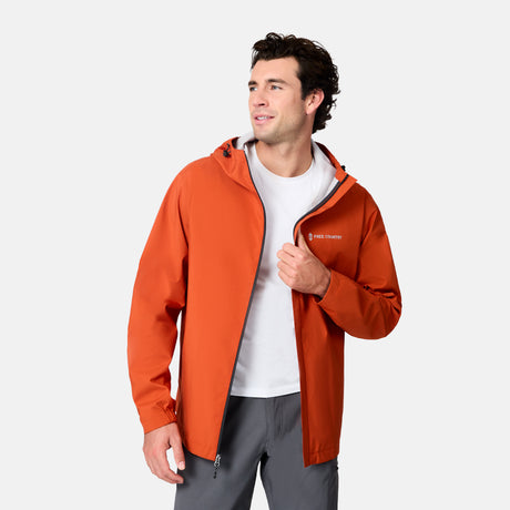 A man smiles to his left while wearing the Free Country Men's Hydro Lite X2O Spectator Waterproof Jacket in bright orange over a white T-shirt and gray pants, standing against a plain light background.