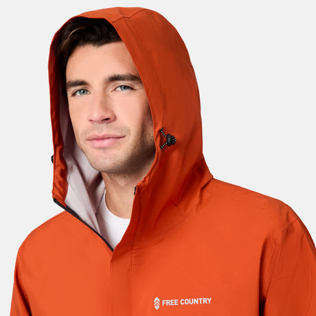 A man wearing a Free Country Men's Hydro Lite X2O Spectator Waterproof Jacket in orange with the hood up looks at the camera. The brand logo appears on the chest against a plain light gray background.