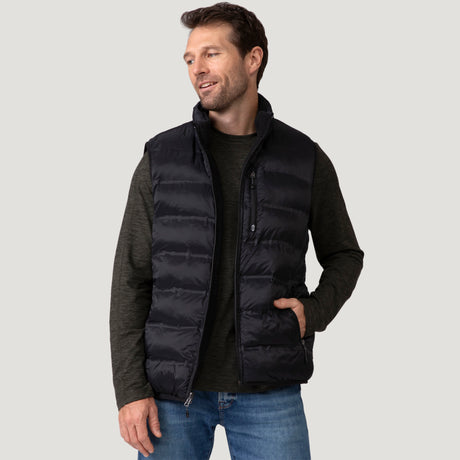[Vince is 6'1" wearing a size Medium.] Man wearing the Free Country Men's Pine Creek Quilted Reversible Vest in black, featuring a quilted outer layer, cozy curly fleece reverse, and REPREVE® synthetic fill, styled with a black long-sleeve shirt and dark jeans.