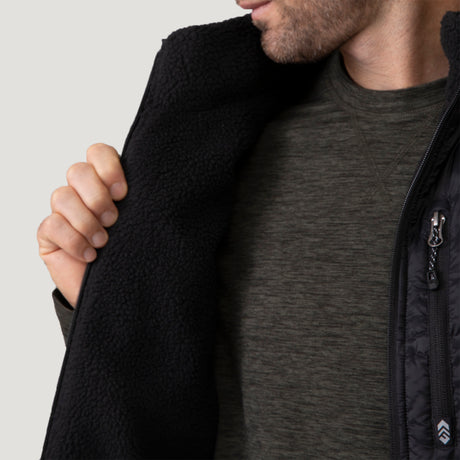 [Vince is 6'1" wearing a size Medium.] Man wearing the Free Country Men's Pine Creek Quilted Reversible Vest in black, featuring a quilted outer layer, cozy curly fleece reverse, and REPREVE® synthetic fill, styled with a black long-sleeve shirt and dark jeans.