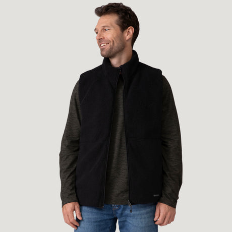 [Vince is 6'1" wearing a size Medium.] Man wearing the Free Country Men's Pine Creek Quilted Reversible Vest in black, featuring a quilted outer layer, cozy curly fleece reverse, and REPREVE® synthetic fill, styled with a black long-sleeve shirt and dark jeans.