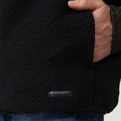 [Vince is 6'1" wearing a size Medium.] Man wearing the Free Country Men's Pine Creek Quilted Reversible Vest in black, featuring a quilted outer layer, cozy curly fleece reverse, and REPREVE® synthetic fill, styled with a black long-sleeve shirt and dark jeans.