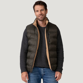 A man wears the Men's Pine Creek Quilted Reversible Vest by Clearance, in dark green with REPREVE fill, over a gray sweater and blue jeans, standing against a light gray background and smiling softly with relaxed hands.