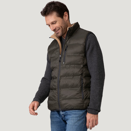 A man wears the Clearance Men's Pine Creek Quilted Reversible Vest with REPREVE fill over a gray knit sweater and blue jeans, smiling and looking down against a plain light background.