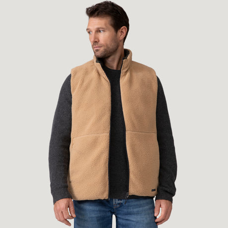 A man models the Clearance Men's Pine Creek Quilted Reversible Vest in tan with REPREVE fill, layered over a dark gray sweater and blue jeans, standing at an angle against a plain light gray background.
