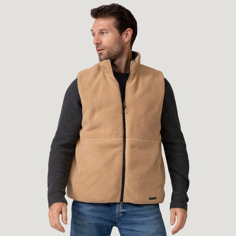 A man models the Clearance Men's Pine Creek Quilted Reversible Vest, featuring REPREVE fill—a tan quilted zip-up over a dark gray sweater and blue jeans—standing slightly turned against a plain light gray background.