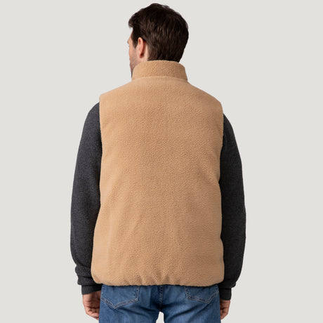 A man is seen from behind wearing the Clearance Men's Pine Creek Quilted Reversible Vest in tan over a dark long-sleeve shirt and blue jeans, standing against a plain light gray background.