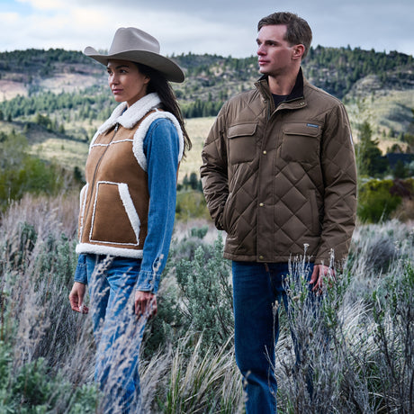 A man and a woman standing outdoors in a grassy field with hills in the background, wearing jackets. The woman wears a brown shearling-lined vest with white trim and a gray cowboy hat, and the man wears a brown diamond-quilted jacket.