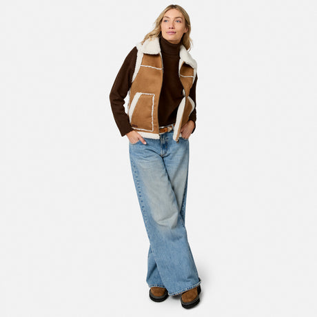 Front view of a woman wearing a tan faux suede shearling reversible vest over a brown turtleneck.