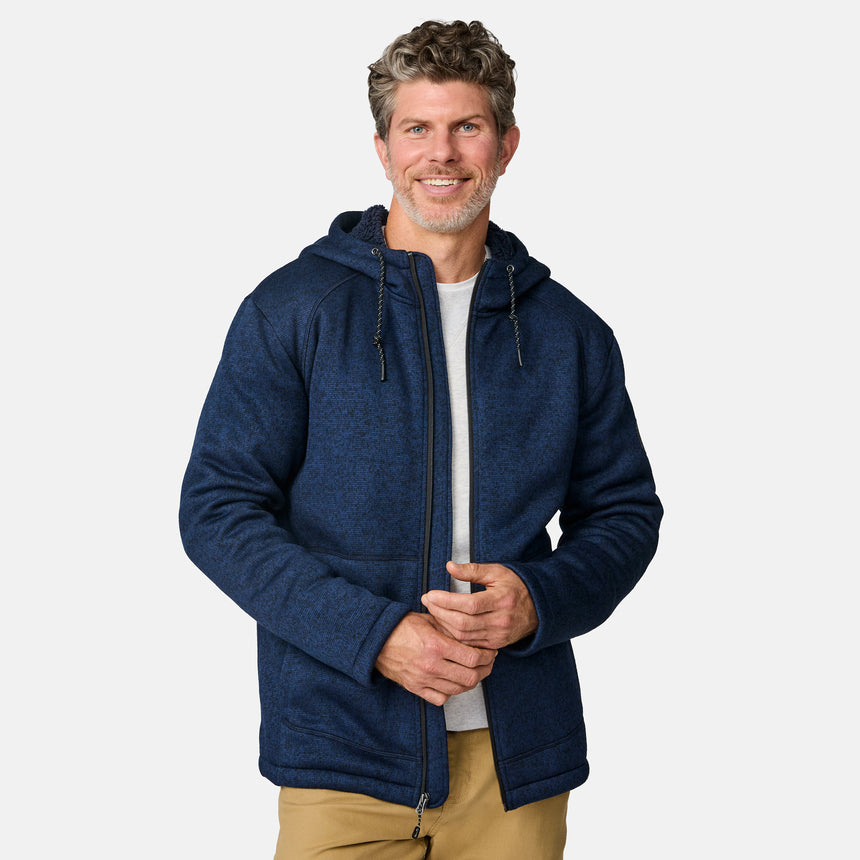 [Kolby is 6’2” wearing a size Medium.] Man wearing the Free Country Men's Northwood Mountain Fleece Hoodie in heather navy with black drawstrings, standing with hands in pockets against a plain, light background.