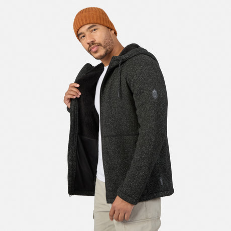 Man wearing a dark Free Country Men’s Mountain Fleece jacket over a white t-shirt and tan pants, with his hand inside the open sherpa hoodie.