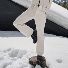 A person wearing cream-colored Free Country Women's Stormlite Super Softshell® Ski Pants and black boots balances on one leg atop a tree stump in the snow, with a snow-covered building and mountain in the background.