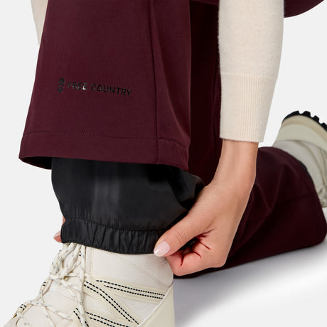 A person in burgundy Free Country Women’s Stormlite Super Softshell® Ski Pants kneels, adjusting the elastic cuff over white boots, paired with a cream sweater for warmth and style.