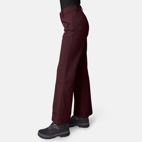 A woman wears Free Country Women’s Stormlite Super Softshell® Ski Pant in burgundy, a black shirt, and black hiking boots, standing on a light gray background. Only the lower half of her body is visible from the side.