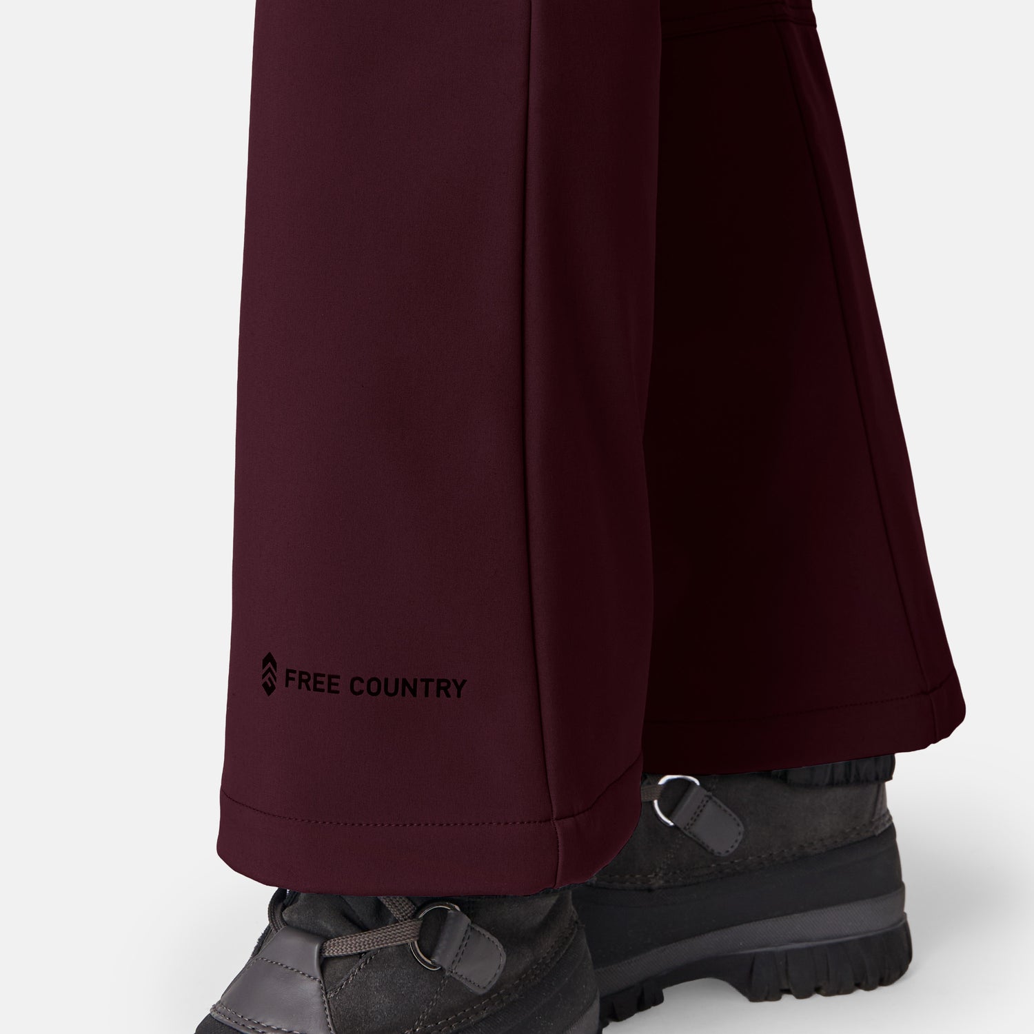 Close-up of a person wearing Free Country Women’s Stormlite Super Softshell® Ski Pant in dark burgundy with water resistance, four-way stretch, and logo on the leg. Gray hiking boots. Plain white background.